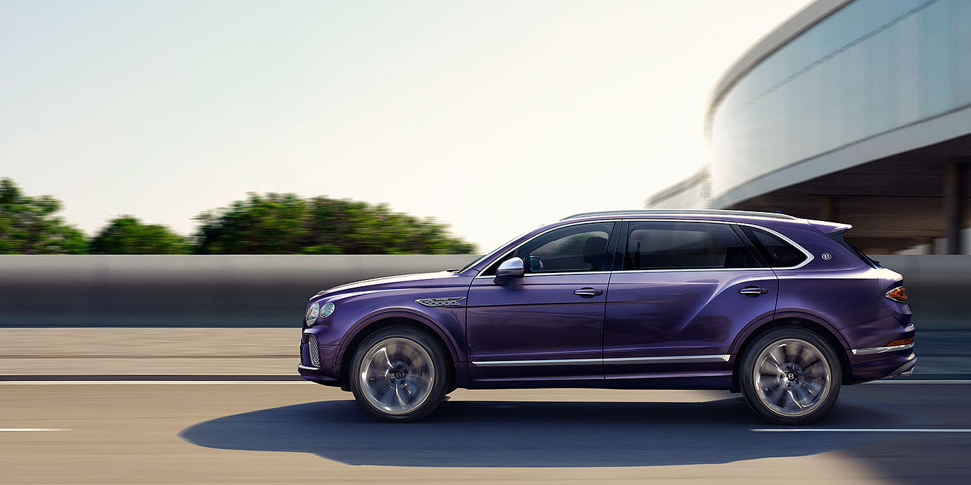 Bentley Beirut Bentley Bentayga Extended Wheelbase Mulliner luxury SUV profile in Tanzanite Purple paint, driving on an country road past trees.