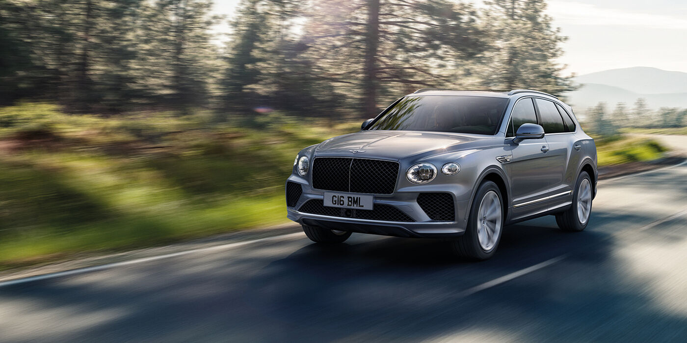Bentley Beirut Bentley Bentayga luxury SUV front three quarter in Moonbeam paint, driving on an country road past trees.